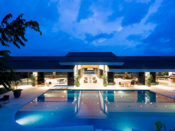 A modern building at night features a large, illuminated swimming pool in front, surrounded by trees and lit by ambient lighting.