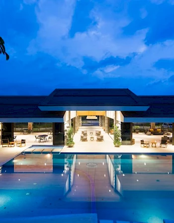 A luxurious modern building with a large illuminated swimming pool in front, set against a twilight sky.