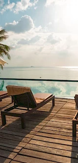 Lounge chairs and umbrellas beside a tranquil pool overlook the ocean, with palm trees and a sunny sky creating a tropical ambiance.