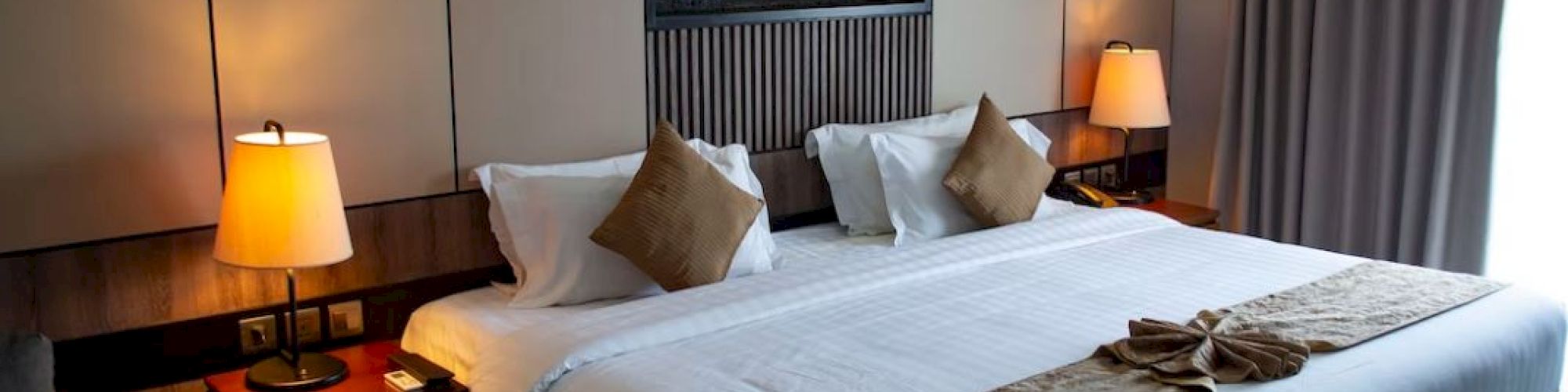 The image shows a neatly arranged hotel room with a large bed, side tables with lamps, and a decorative wall piece above the bed.