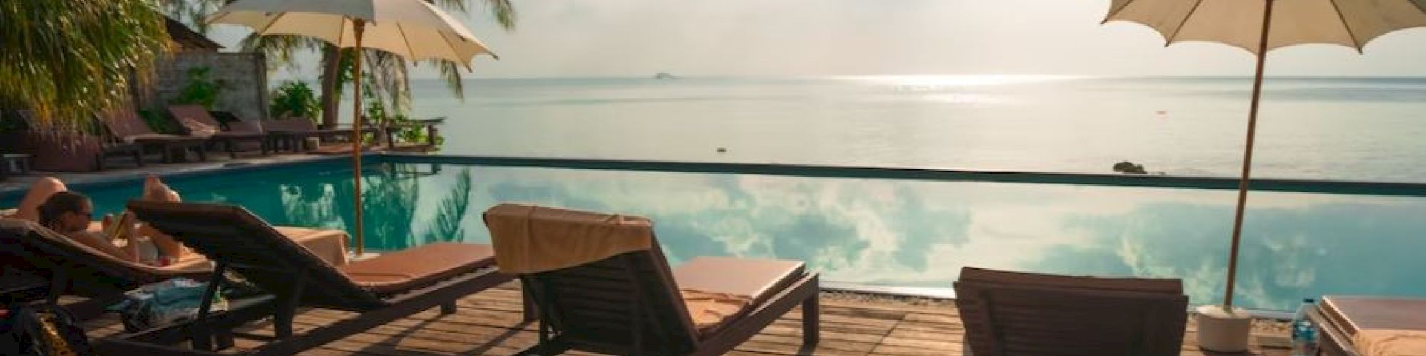 Poolside view with lounge chairs and umbrellas on a wooden deck, overlooking a calm sea under a partly cloudy sky.