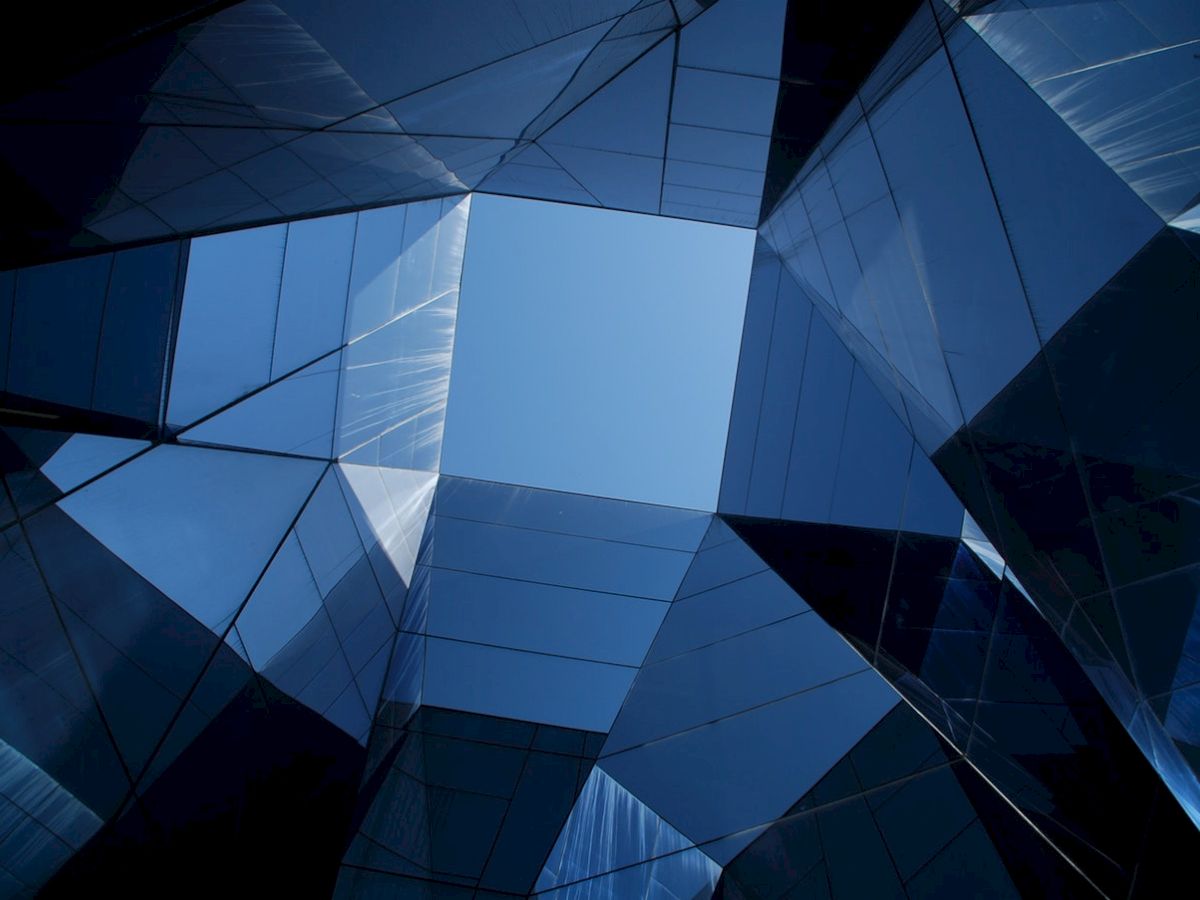 The image shows a geometric view of buildings with reflective glass surfaces, creating a kaleidoscopic effect against a clear blue sky.
