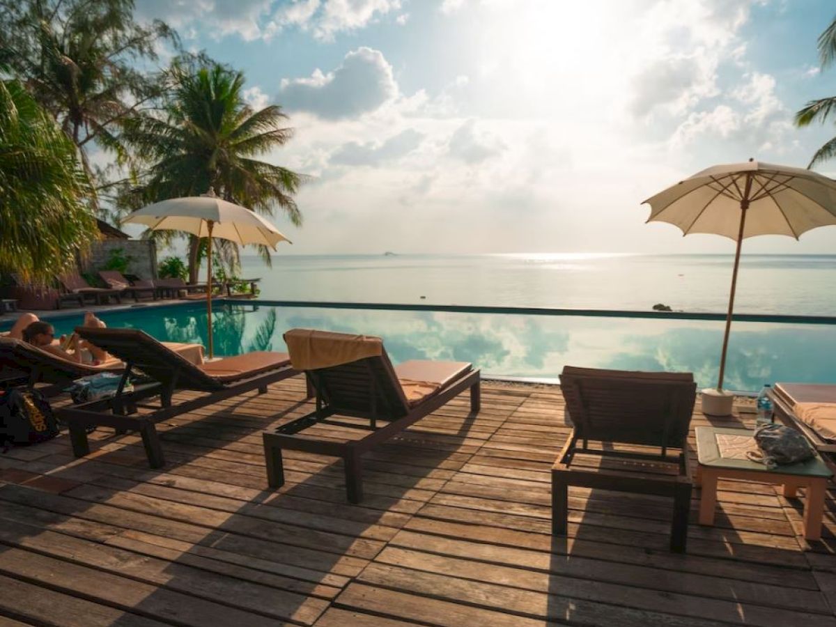 A serene poolside scene with lounge chairs, umbrellas, and palm trees overlooking a calm body of water under a partly cloudy sky.