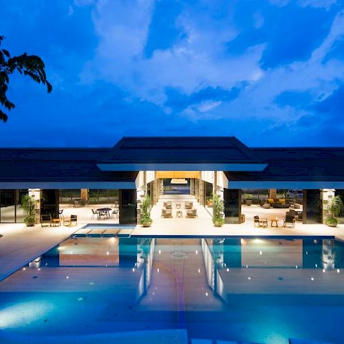 A luxurious modern building at night with a well-lit swimming pool reflecting the structure, set against a dramatic evening sky.