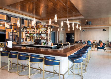 A modern bar with sleek design, blue cushioned stools, hanging lights, and bottles on shelves. Cozy seating area in the background.