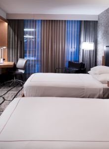 The image shows a modern hotel room with two beds, a wall-mounted TV, a desk, lamps, and curtains.