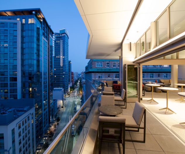 A modern rooftop balcony with seating overlooks a city street and tall glass buildings at dusk, creating a serene urban atmosphere.