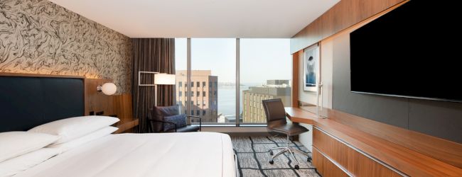 A modern hotel room with a large bed, a TV, a desk, and a chair, overlooking a city view through a big window.