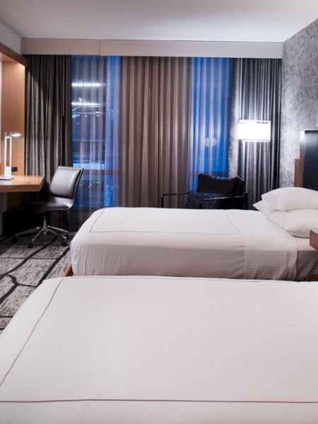 The image shows a modern hotel room with two double beds, a TV on the wall, a desk, and a window with curtains.