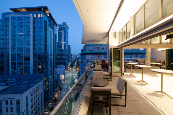A modern rooftop terrace with seating overlooks a city street and tall, illuminated buildings, creating a vibrant urban evening scene.