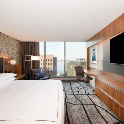 A modern hotel room with a large bed, a desk, a wall-mounted TV, and a window offering a city view.