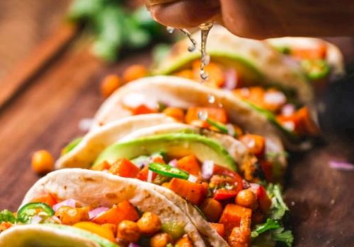 Tacos filled with chickpeas, avocado slices, and veggies are arranged on a board. A hand is squeezing lime juice over them.