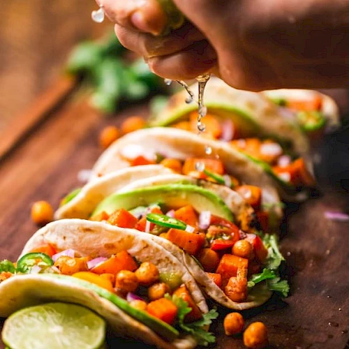 Tacos filled with chickpeas, avocado slices, and veggies are arranged on a board. A hand is squeezing lime juice over them.