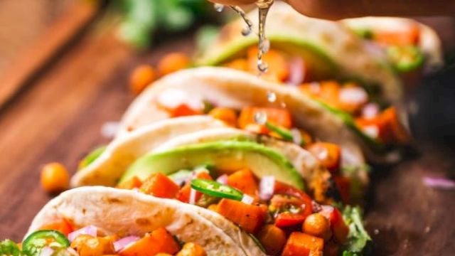 Tacos filled with chickpeas, avocado slices, and veggies are arranged on a board. A hand is squeezing lime juice over them.