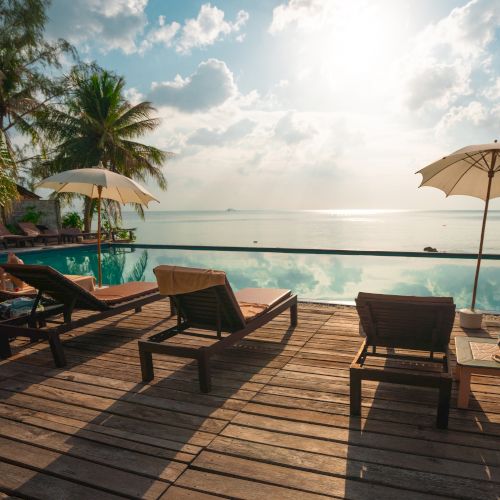 A tranquil poolside scene with deck chairs, umbrellas, palm trees, and a stunning ocean view under a bright sunlit sky.