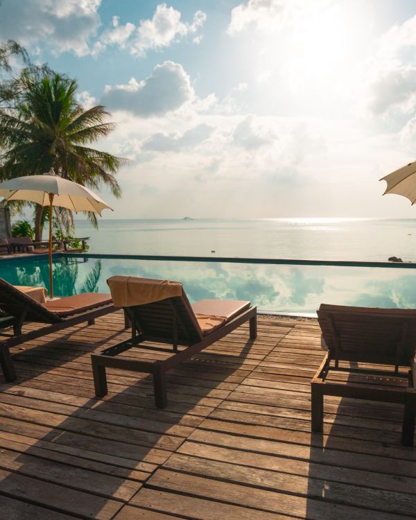 A tranquil poolside scene with deck chairs, umbrellas, palm trees, and a stunning ocean view under a bright sunlit sky.