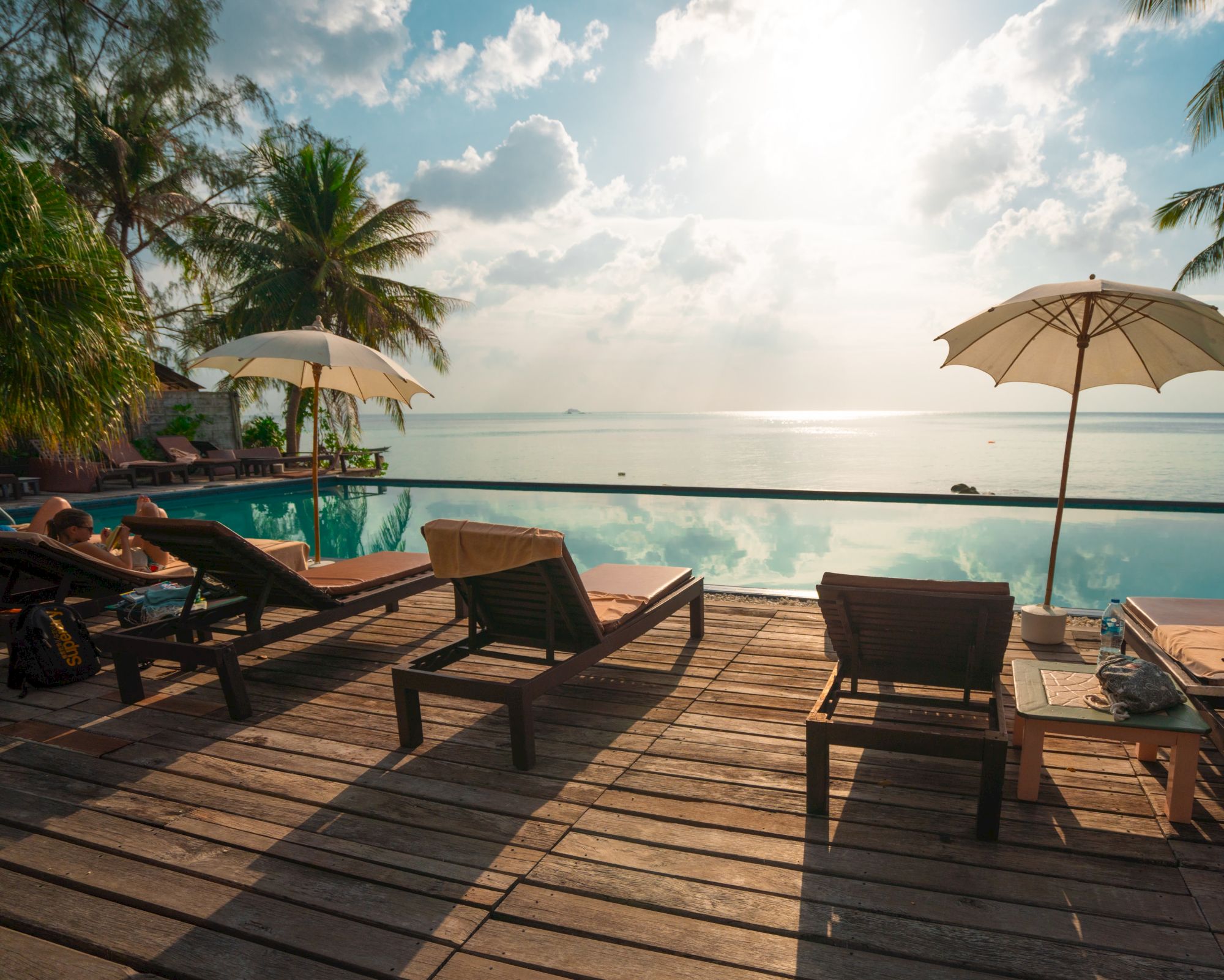 A serene poolside scene with lounge chairs, umbrellas, and palm trees overlooking a calm ocean under a bright sky.