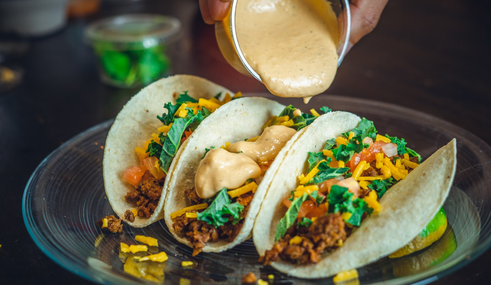 Three tacos with ground meat, cheese, lettuce, and sauce being poured over them.