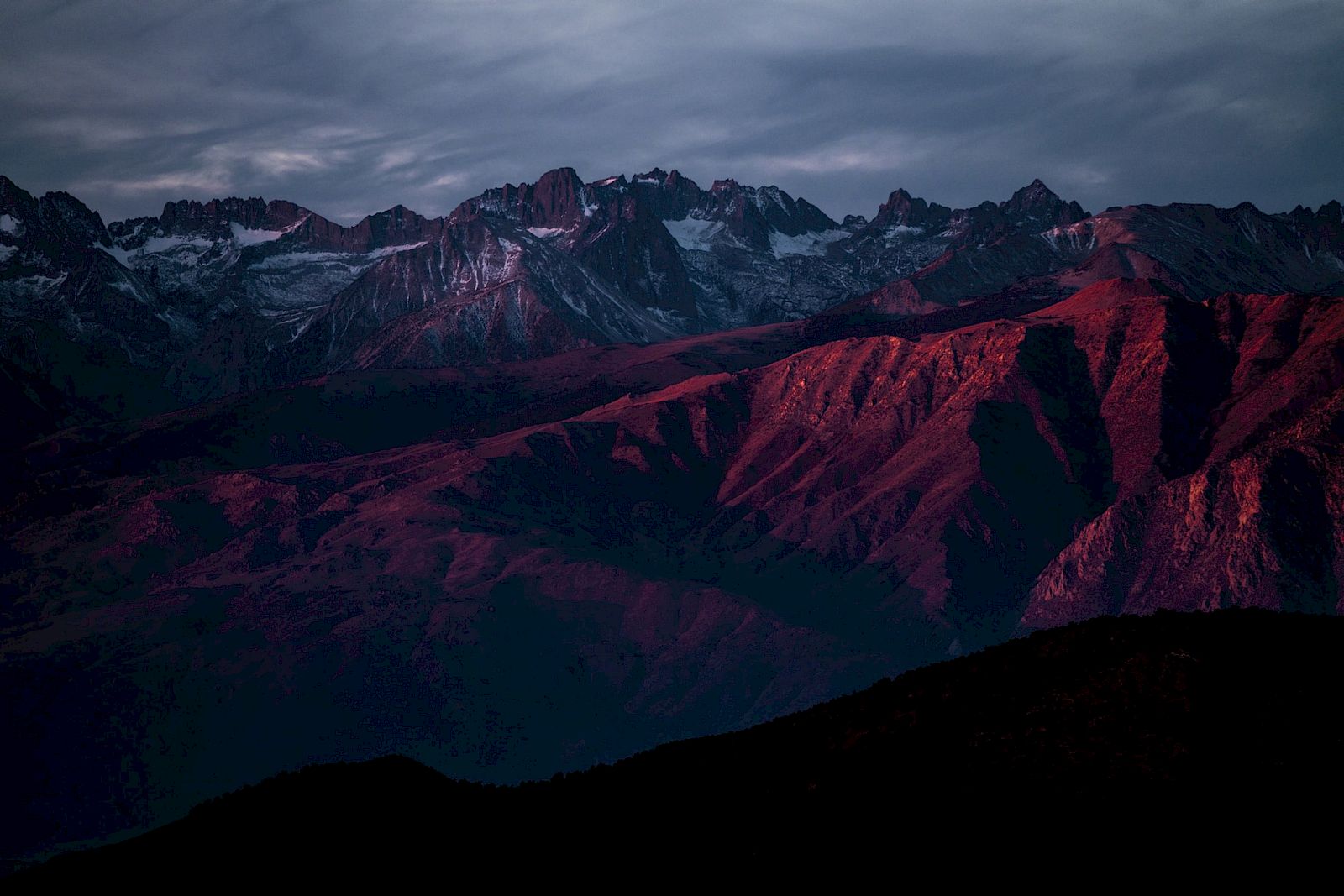 A mountain range lit by reddish-purple light under a cloudy sky, creating a dramatic and moody landscape scene.