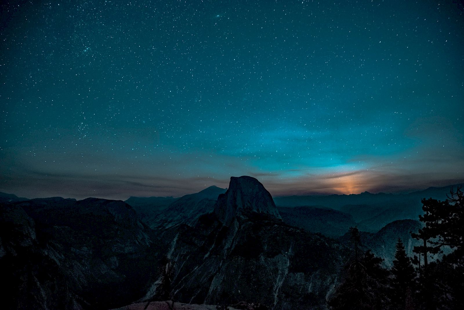 A night landscape with star-filled sky over mountains, featuring a prominent peak and silhouetted trees.