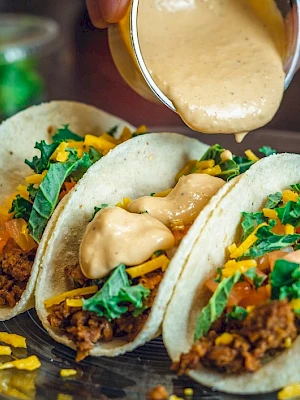 Three tacos filled with ground meat, cheese, lettuce, and tomato, with sauce being poured over them on a plate.