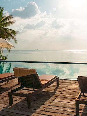 A serene poolside view with lounge chairs, umbrellas, and palm trees overlooking a calm ocean under a bright sky.