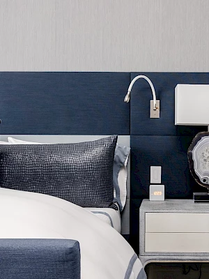 A modern bedroom featuring a bed with blue and gray pillows, a blue headboard, and a nightstand with a lamp and a wall light.