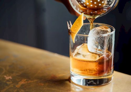 A drink is being poured into a glass with ice and a twist of citrus peel on a wooden surface.