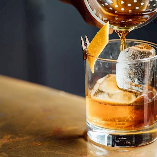 A drink is being poured into a glass with ice and a twist of citrus peel on a wooden surface.