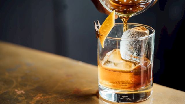 A drink is being poured into a glass with ice and a twist of citrus peel on a wooden surface.