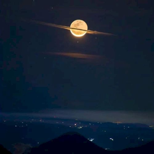The image shows a moon with thin clouds resembling Saturn's rings, above distant hills and city lights in the night.