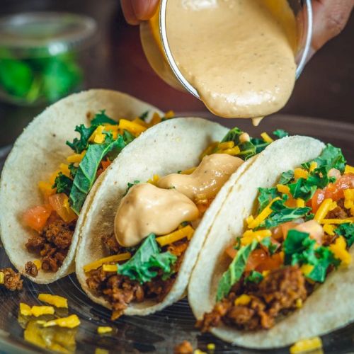 Three tacos filled with ground meat, cheese, greens, and tomatoes, being topped with a creamy sauce on a plate.
