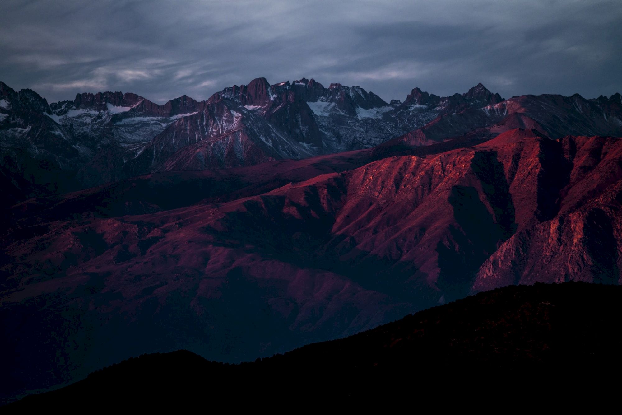 A mountain range is bathed in soft red light, with dramatic clouds overhead, creating a moody landscape scene.