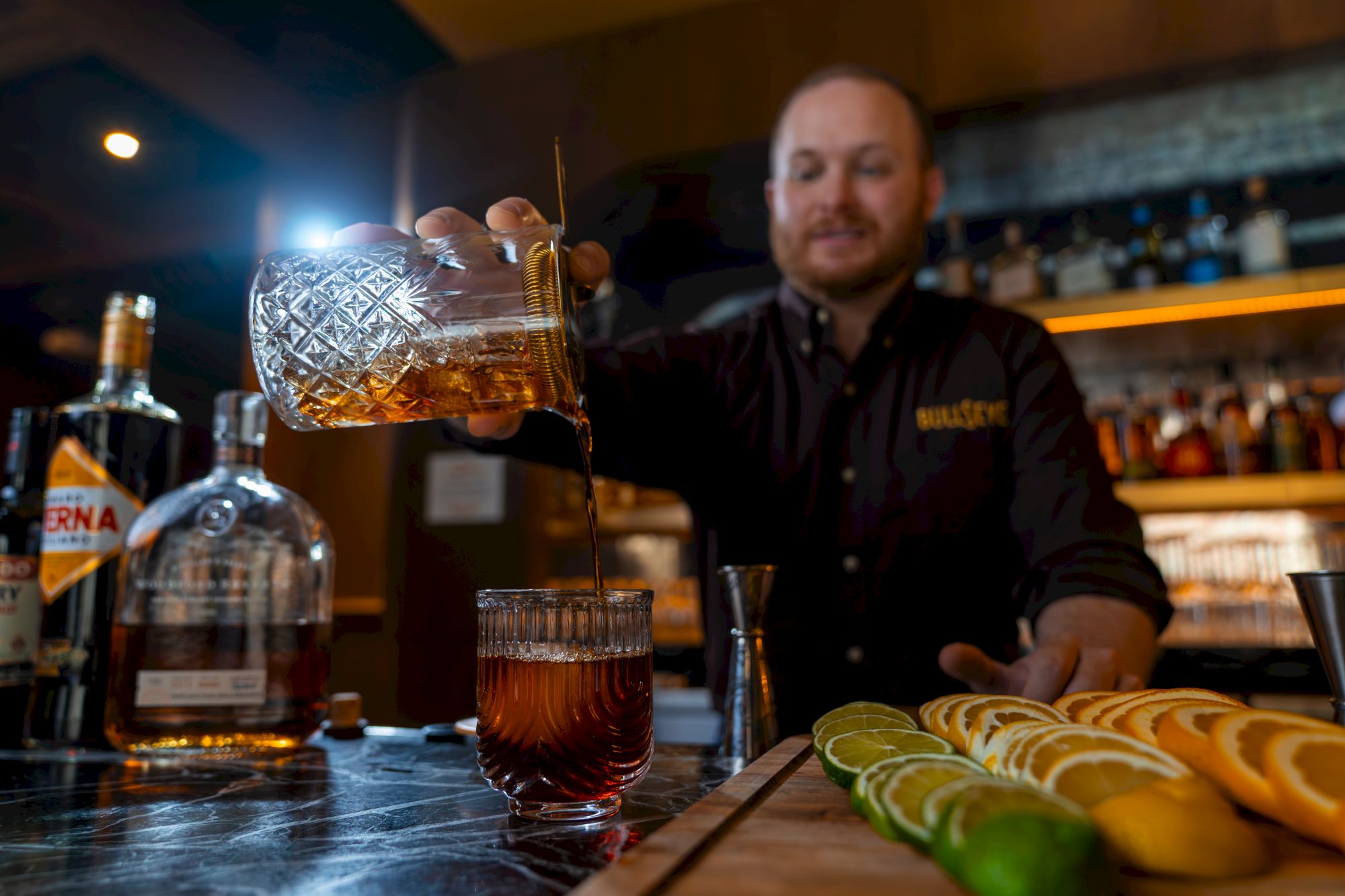 A bartender pours whiskey into a glass at a bar, with a bottle and lime slices on the counter.