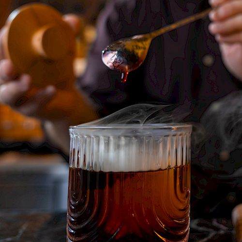 A glass of dark whiskey on the rocks being stirred, with a person in the background using a dropper&mdash;top it at 140 characters, always ending the sentence.