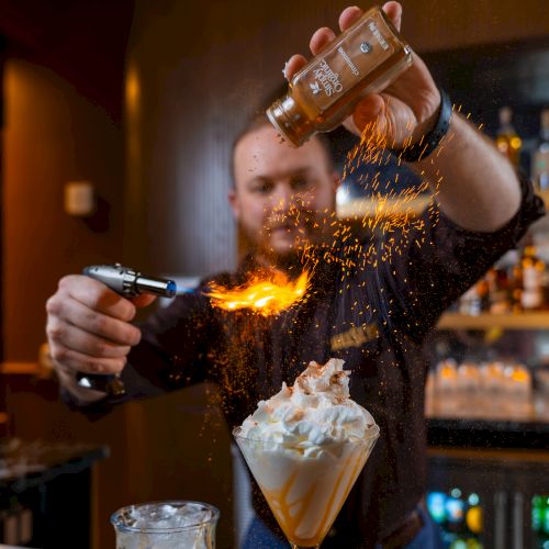 A bartender flames a drink: lighting a flame from a bottle while pouring, with whipped cream-topped beverage in the foreground.