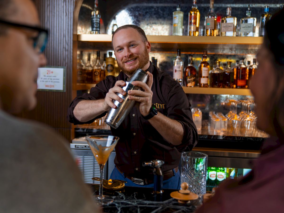 A bartender shakes a cocktail shaker behind the bar while two patrons sit nearby, with bottles and glasses lining the shelves in the background.