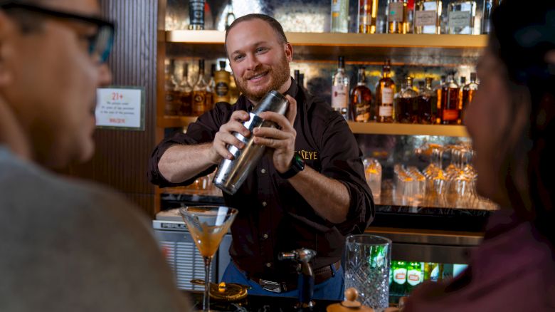 A bartender shakes a cocktail shaker behind the bar while two patrons sit nearby, with bottles and glasses lining the shelves in the background.