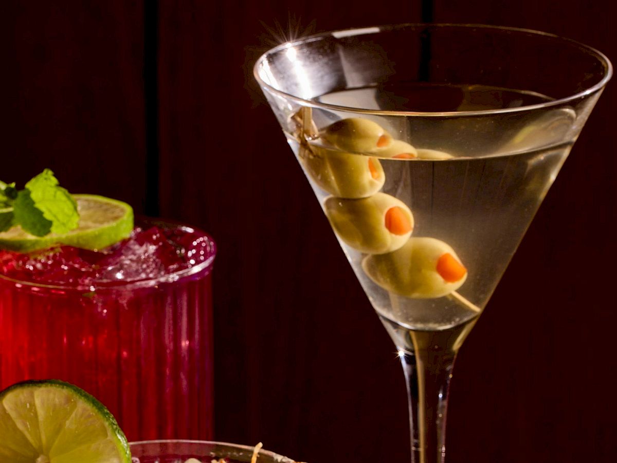 A colorful assortment of cocktails on a dark bar: a red drink with mint and lime, a creamy frosted glass, a pinkish-orange drink with a lemon twist, and a martini with alternating olives, all against a dark wooden backdrop.