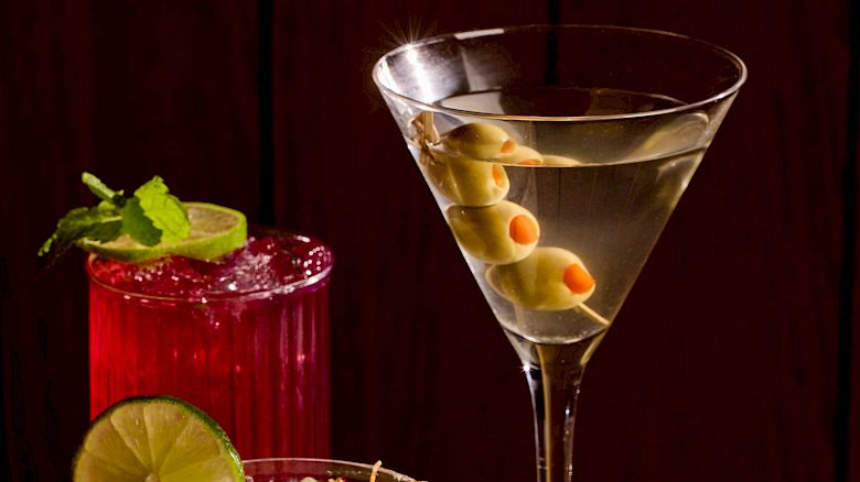 A colorful assortment of cocktails on a dark bar: a red drink with mint and lime, a creamy frosted glass, a pinkish-orange drink with a lemon twist, and a martini with alternating olives, all against a dark wooden backdrop.