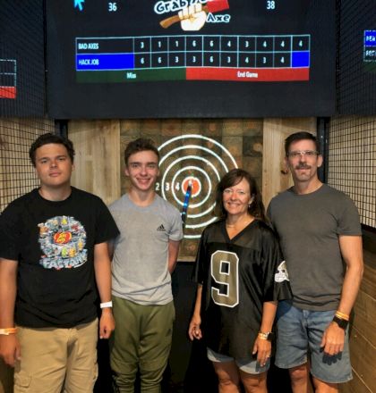 A group of four people standing in front of an axe-throwing target, under a scoreboard displaying scores for "Bad Axes" and "Hack Job."