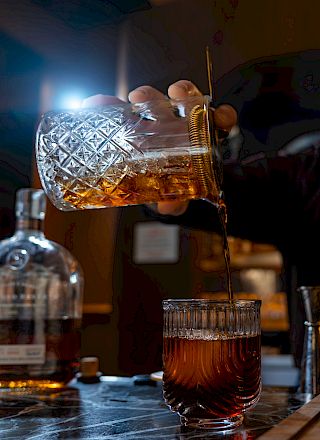 A bartender pours a whiskey or dark liquor over ice in a glass, with a bottle, lime slices, and garnish on the bar.