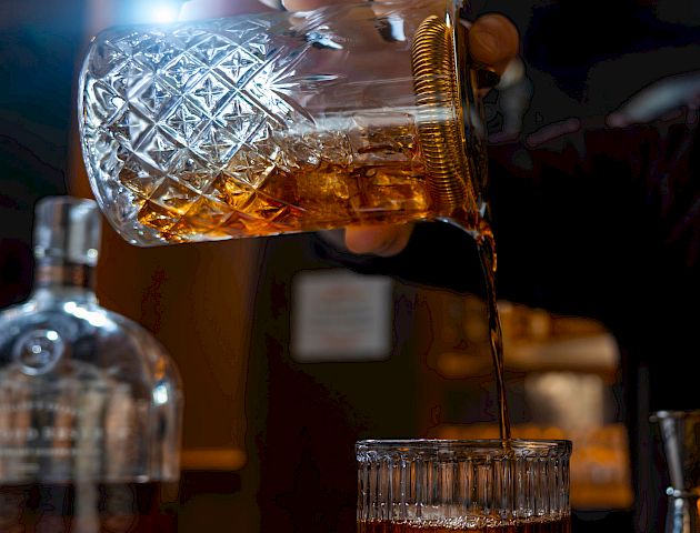 Two beer glasses clink over a dark bar, foamy top and amber liquid; warm lighting and a blurred bottle in the background.