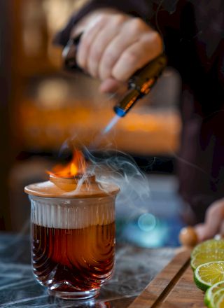 A hand holds a flame torch over a glass mug of dark beer, with lime wedges and skewers nearby; smoking and flame create a dramatic bar scene.