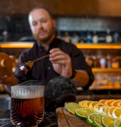 A bartender pours a dark drink from a spoon, with lime and orange slices lined up on the bar in the foreground.