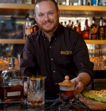 A friendly bartender in a Bullseye shirt behind a marble bar, crafting a drink with a glass, decanter, and lime wedges on display.