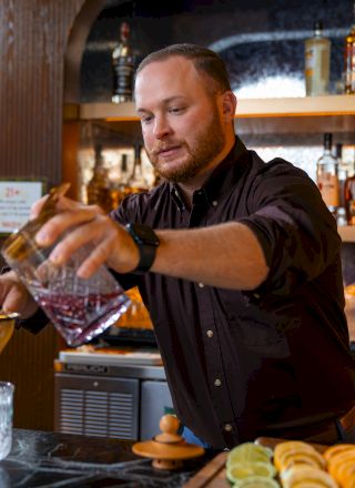 A bartender pouring a drink behind a bar with bottles; a man in a black shirt points forward. Top it at 140 characters, always ending the sentence.