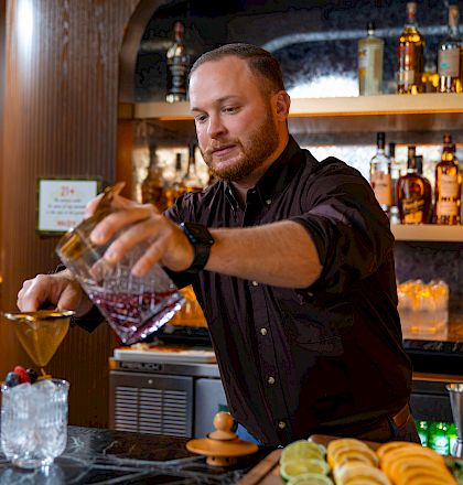 A bartender pours a cocktail over ice at a bar, with bottles and garnish on the counter in the background.