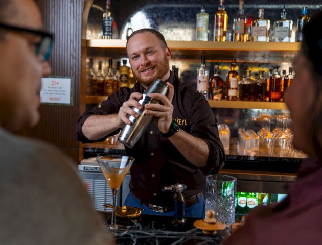 A bartender shakes a cocktail shaker behind a stylish bar, while two patrons watch; bottles line the backlit shelves.