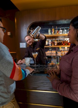 Bartender pours a drink while two customers watch over a dimly lit bar. A lively scene with cocktail whisk and smiles.